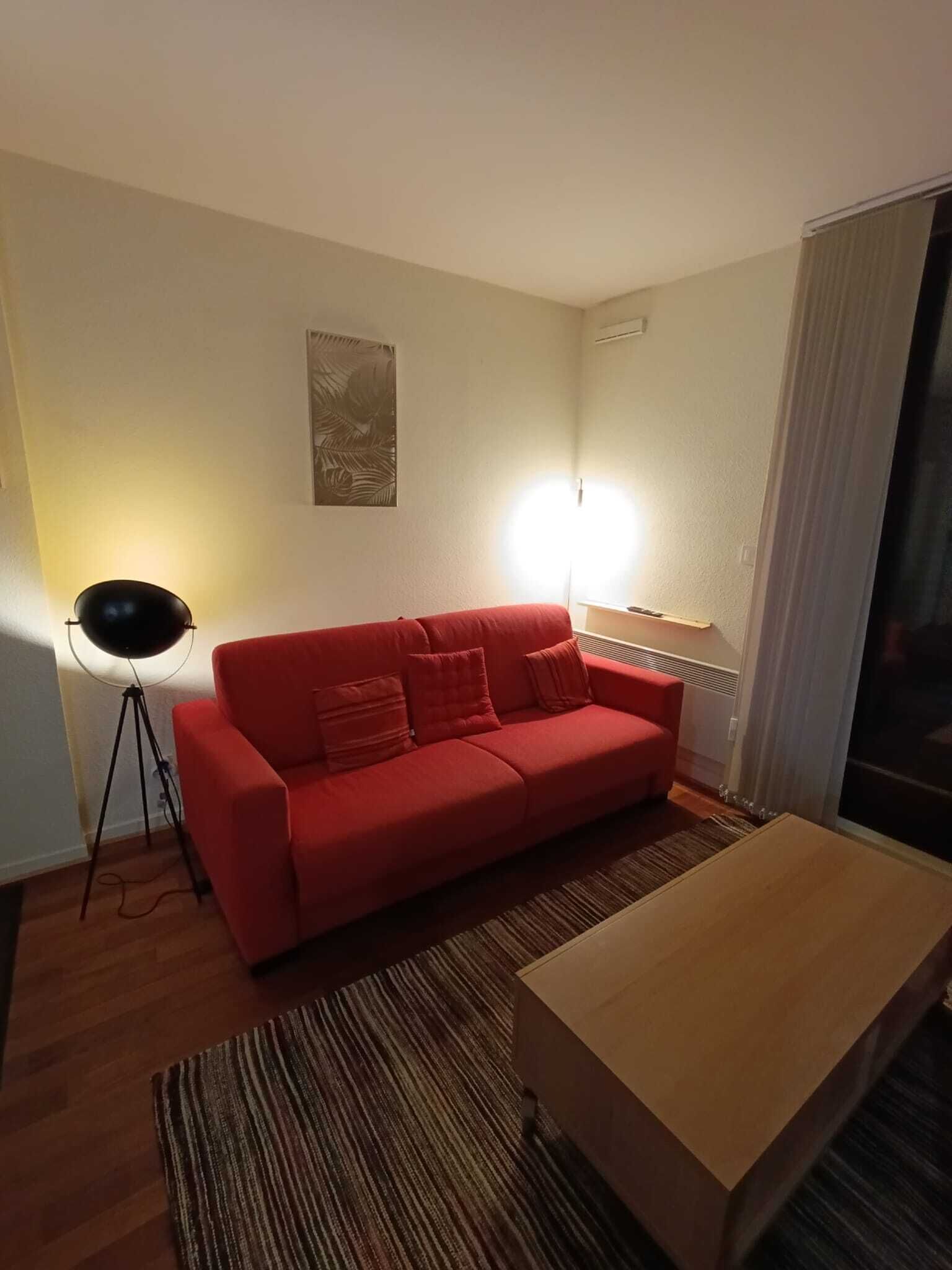 A red sofa, a small wooden table, a striped rug, and a floor lamp in a room with a plain wall and a window with curtains.