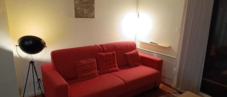 A red sofa, a small wooden table, a striped rug, and a floor lamp in a room with a plain wall and a window with curtains.