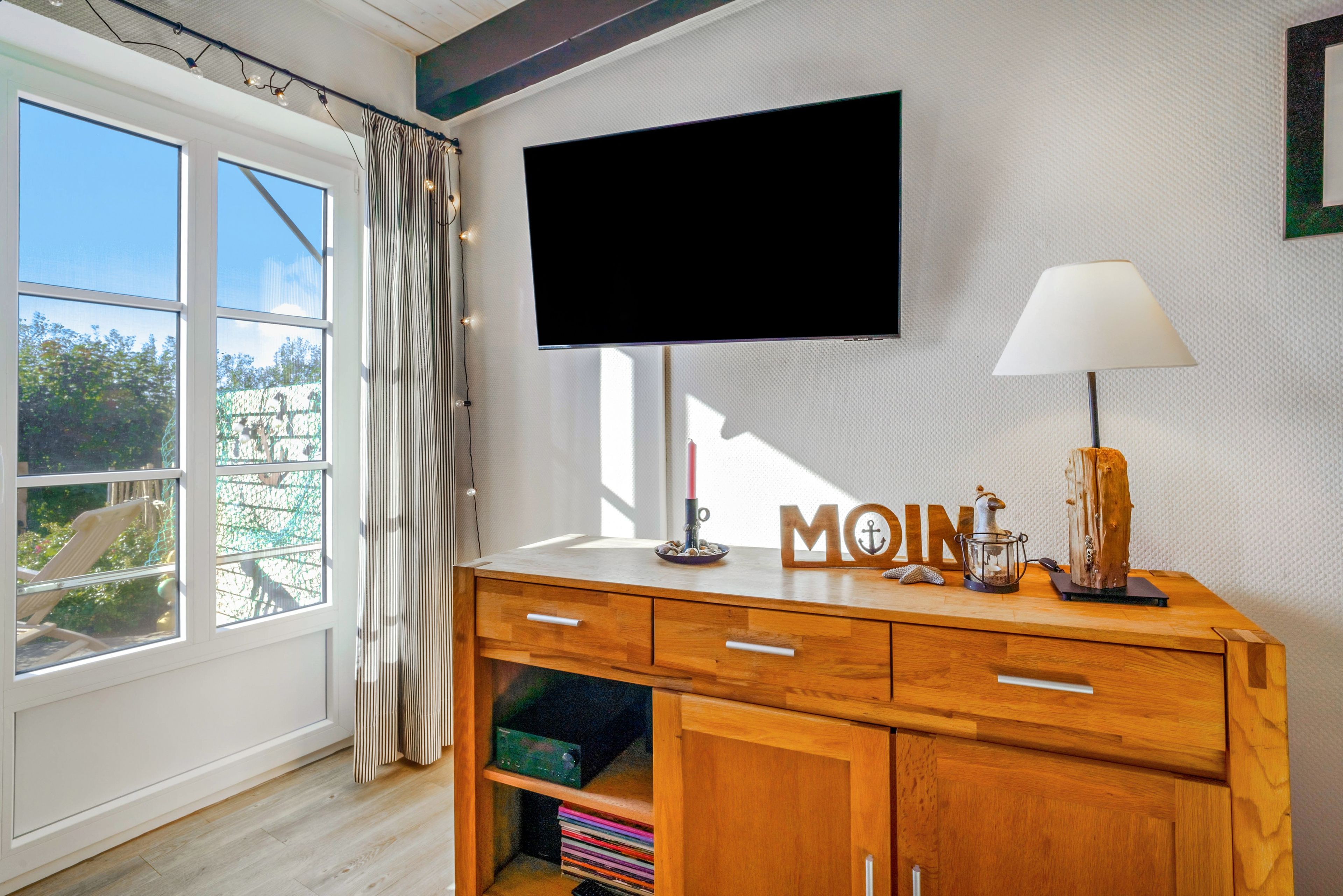 A room with a wooden cabinet, a flat-screen TV mounted on the wall, a lamp, and a window with curtains.