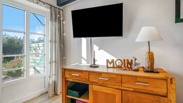 A room with a wooden cabinet, a flat-screen TV mounted on the wall, a lamp, and a window with curtains.