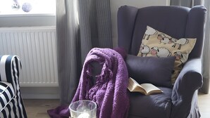 Ein Wohnzimmer mit einem dunklen Sessel, einem gestreiften Stuhl, einer violetten Decke, einem Buch und einer Kerze auf einem Tisch.