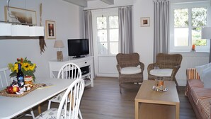 A living room with a television, wicker chairs, a sofa, a dining table with a tray of food, and a window with curtains. - Apartment 'Sonnenkuss' with Private Terrace, Shared Garden & Wi-Fi (Pellworm)