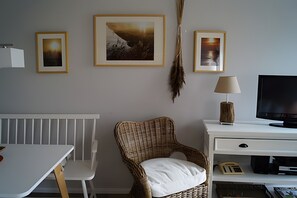 Ein Zimmer mit weißem Schreibtisch, einem Korbstuhl, einem Fernseher und gerahmten Bildern an der Wand.