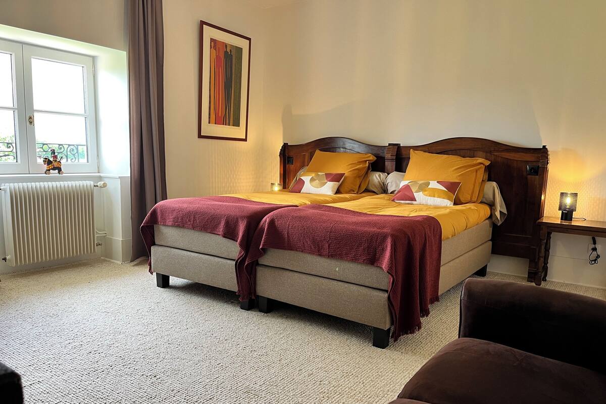 A bedroom with a bed, a radiator, a window, a picture on the wall, and a nightstand with a lamp.