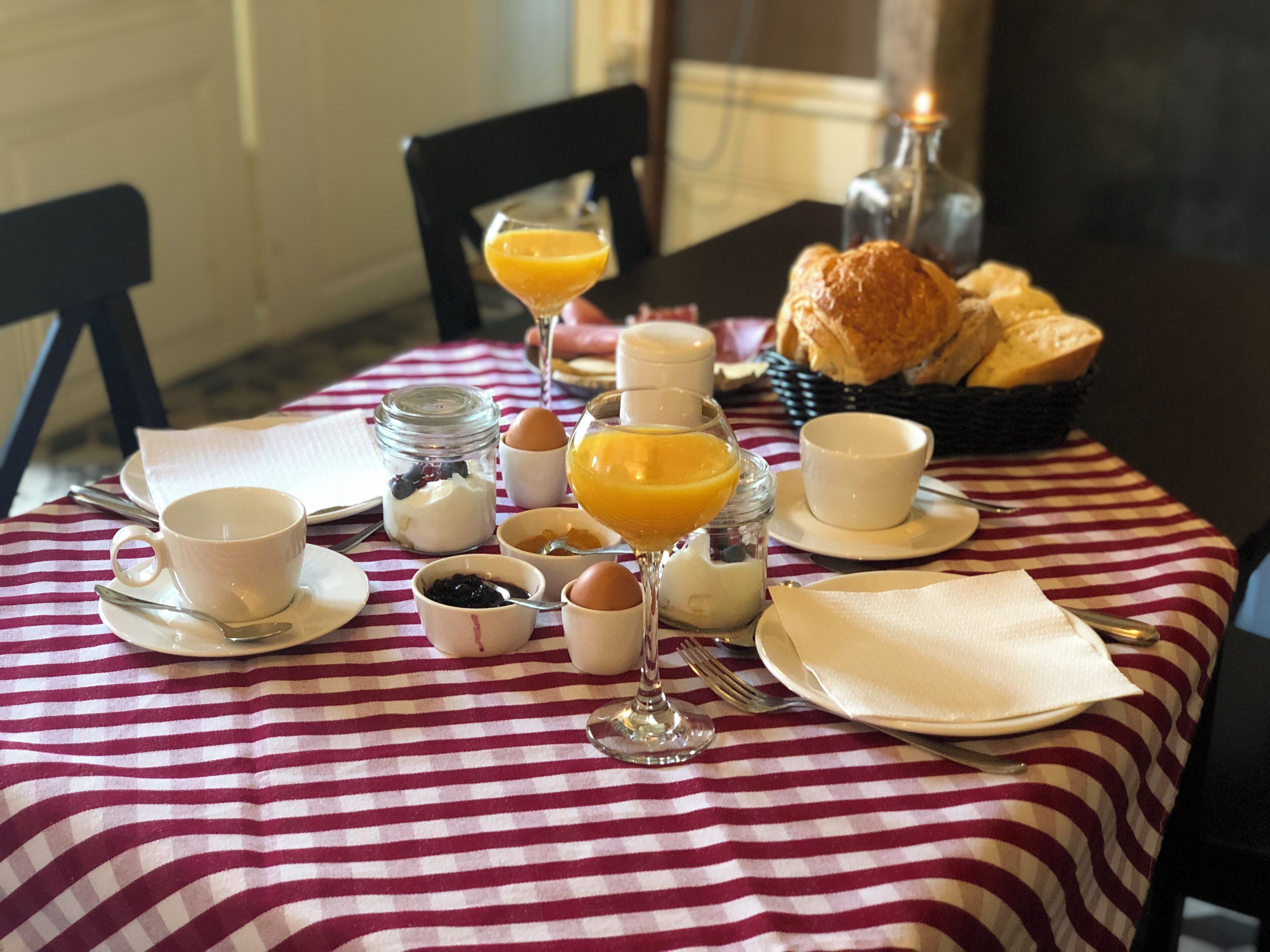 Ein Frühstückstisch mit gestreifter Tischdecke, einem Korb mit Croissants und verschiedenen Frühstücksangeboten, darunter Eier, Toast und Orangensaft.