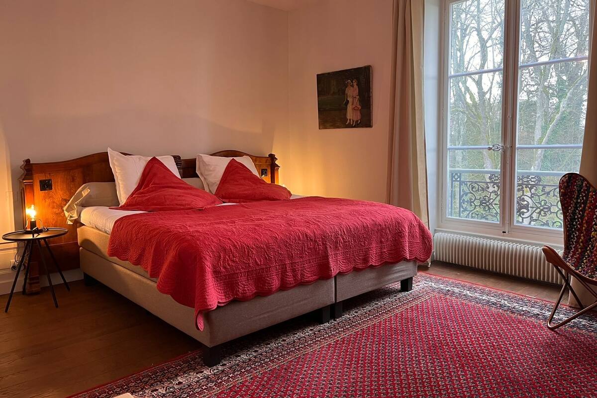 A bedroom with a bed, a nightstand, a chair, and a window with a view of trees.
