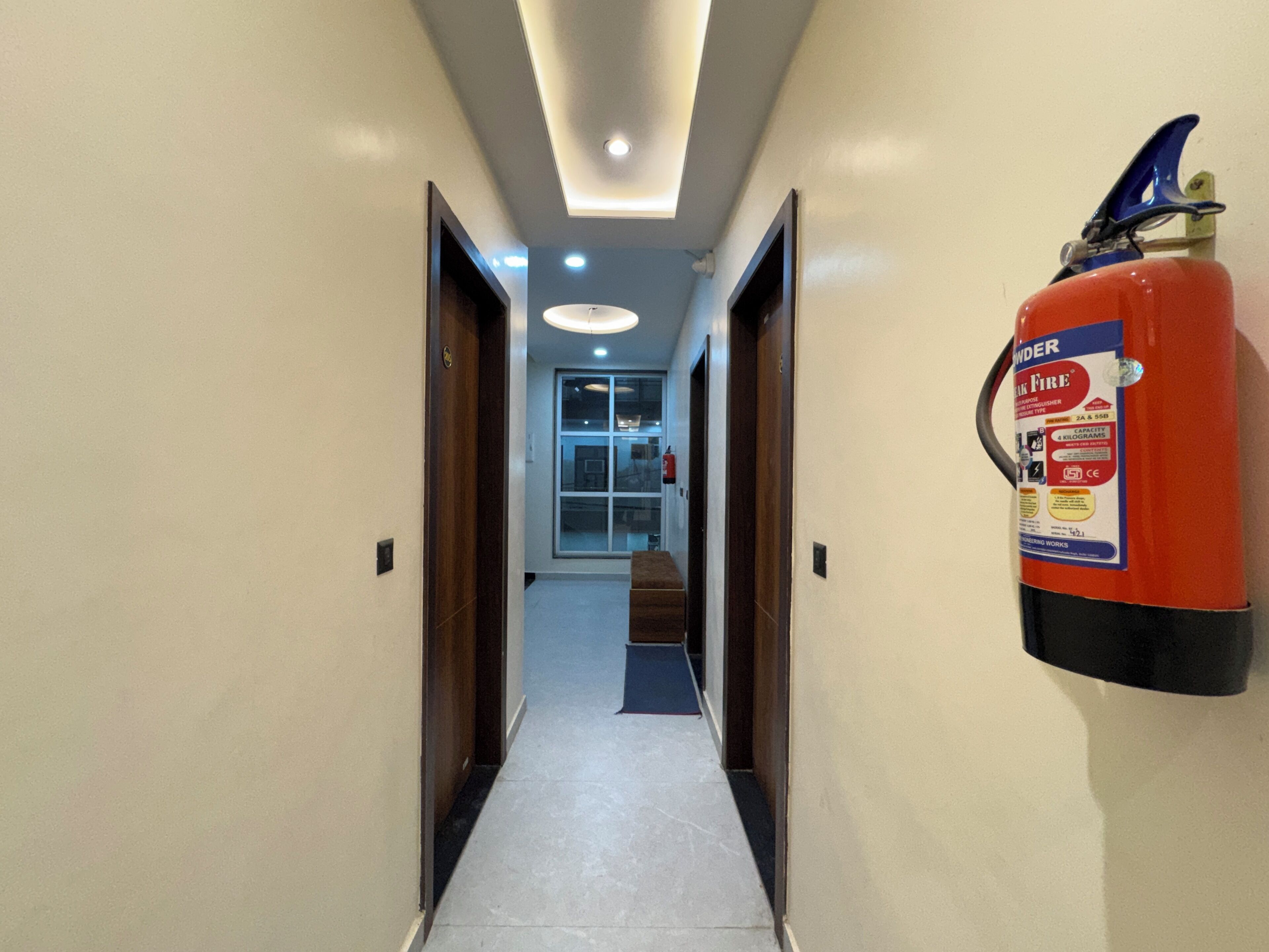 A hallway with a fire extinguisher mounted on the wall, a ceiling light, and a door at the end.