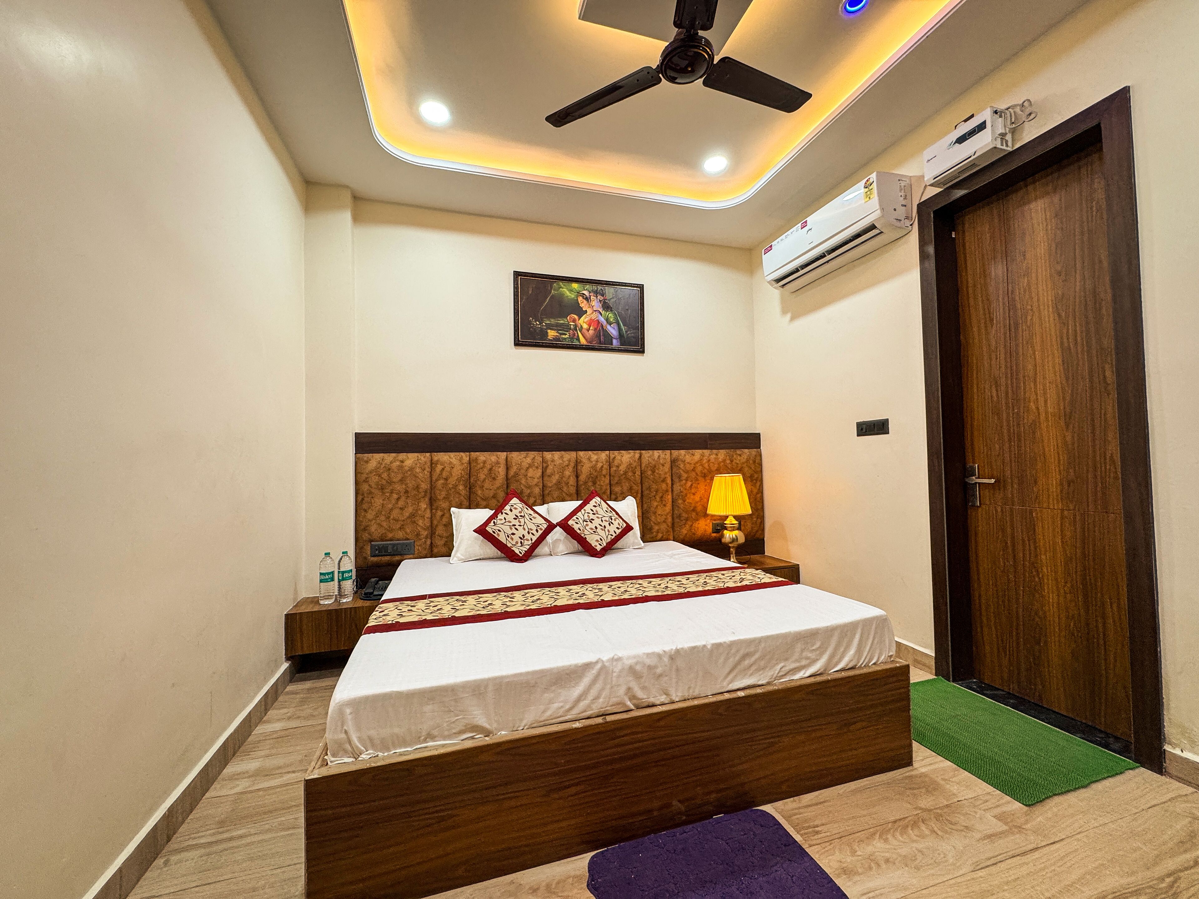 Ein Hotelzimmer mit Bett, Nachttisch, Wand-Klimaanlage, Deckenventilator und einer Tür.