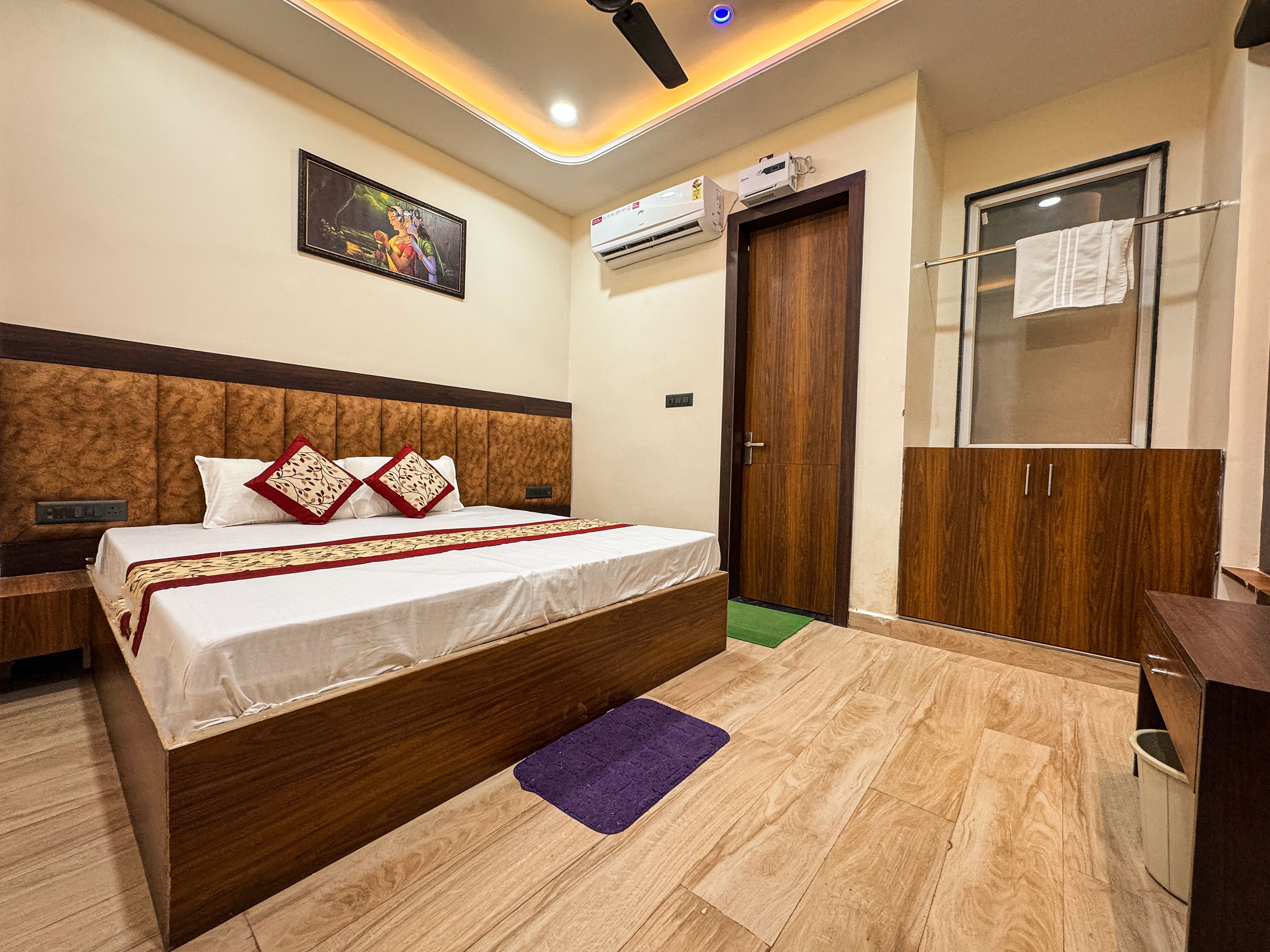 A hotel room with a large bed, a wooden headboard, a ceiling fan, an air conditioning unit, a wardrobe, and a painting on the wall.