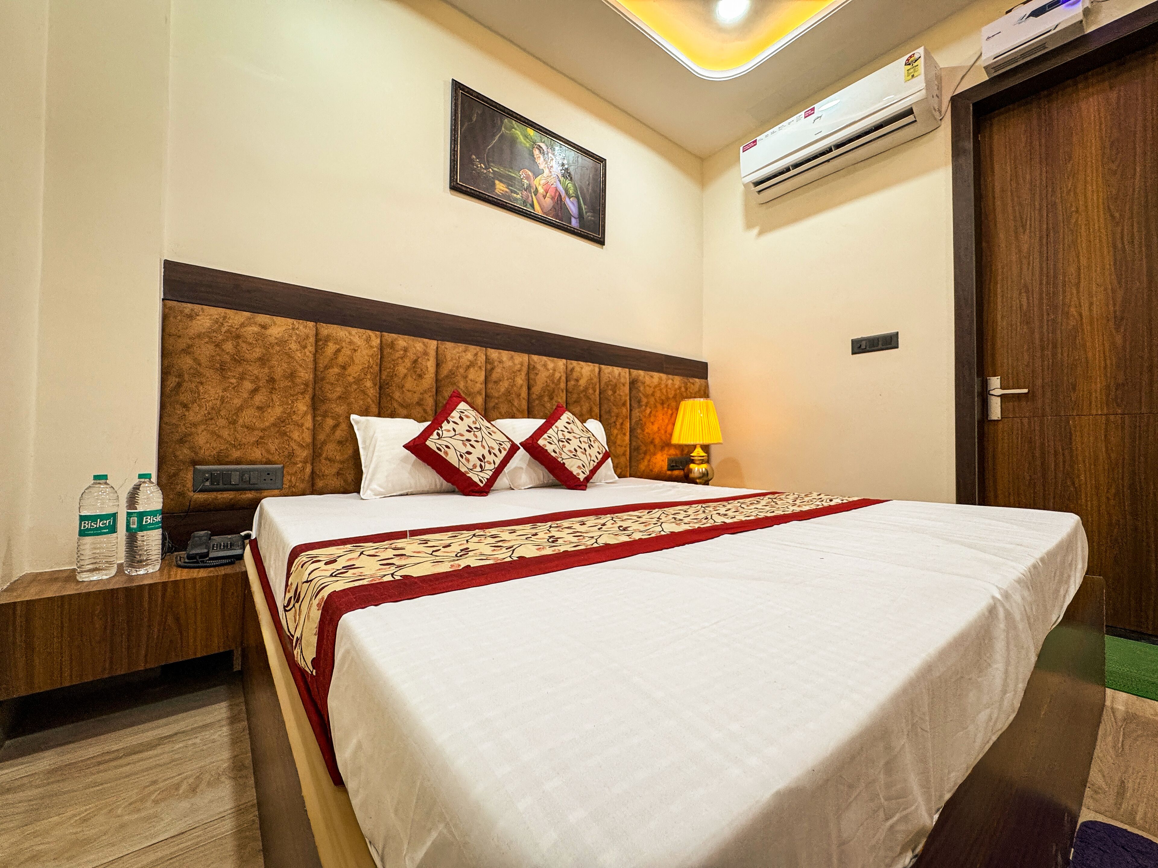 A hotel room with a large bed, a nightstand with water bottles, a lamp, and an air conditioning unit on the wall.