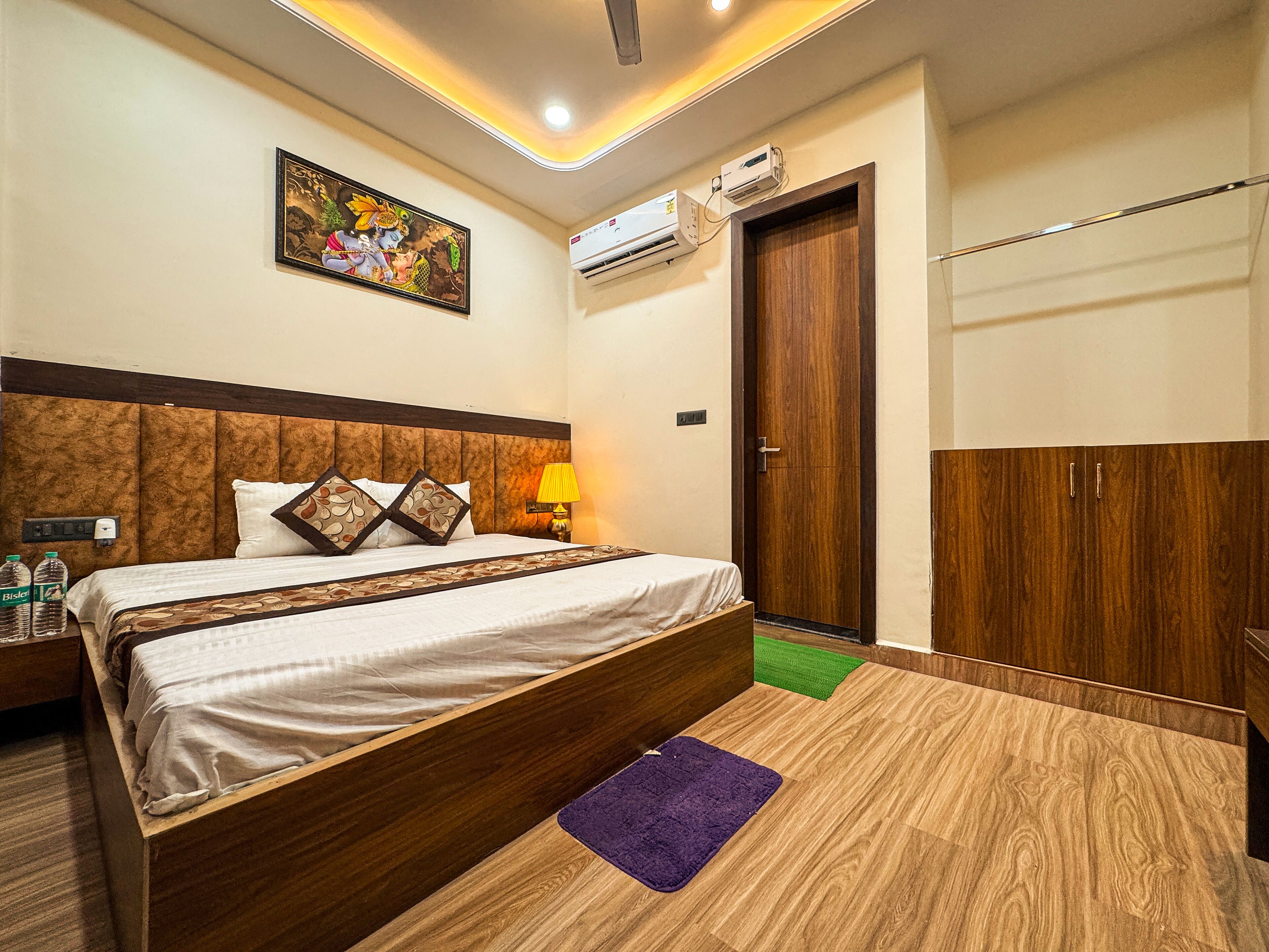 A hotel room with a bed, a nightstand, a lamp, an air conditioning unit, a wardrobe, and a wall-mounted picture.