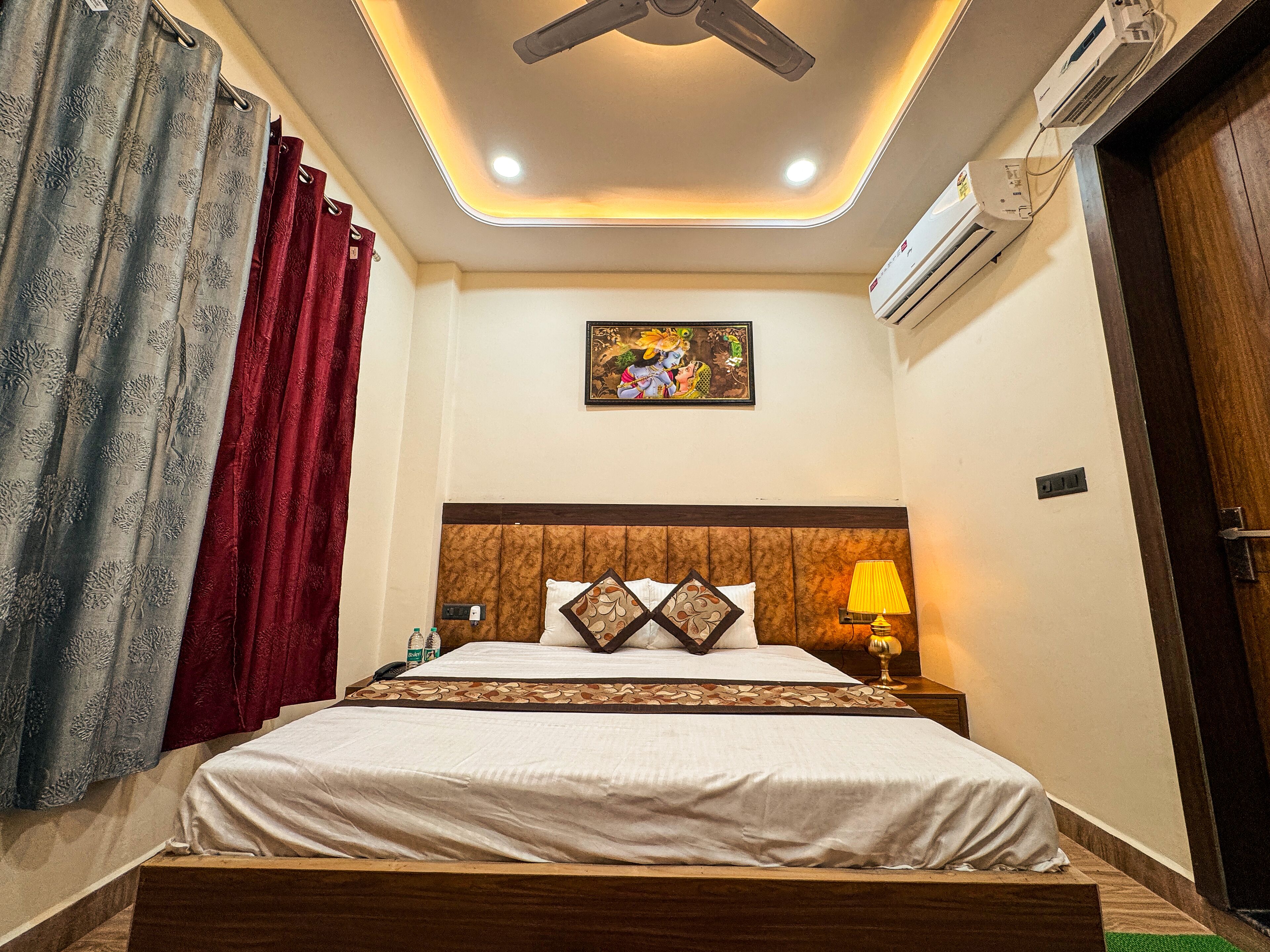 A hotel room with a bed, a bedside table with a lamp, an air conditioning unit, and a ceiling fan.