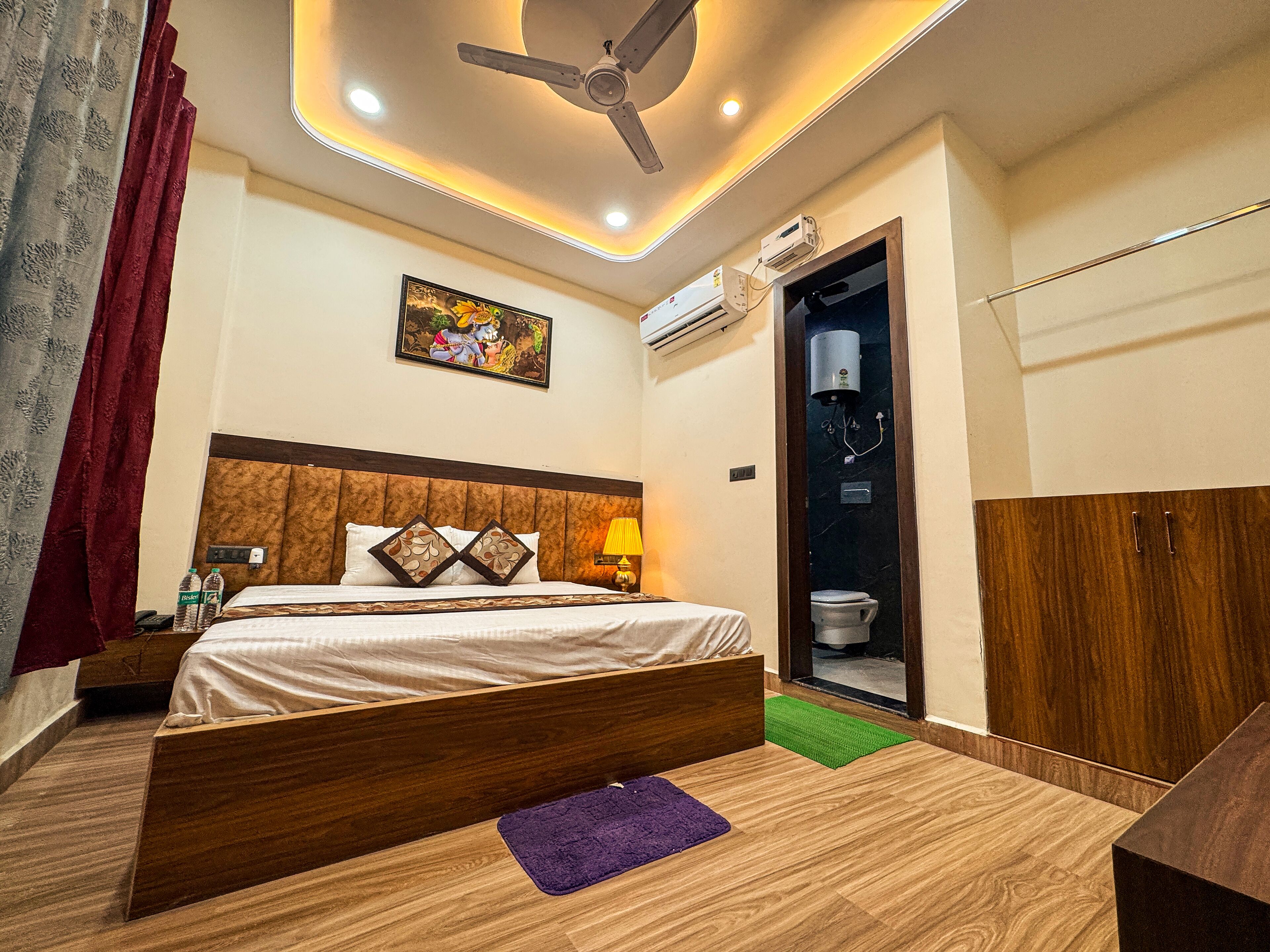 A hotel room with a bed, a ceiling fan, an air conditioning unit, a toilet, and a small green rug.