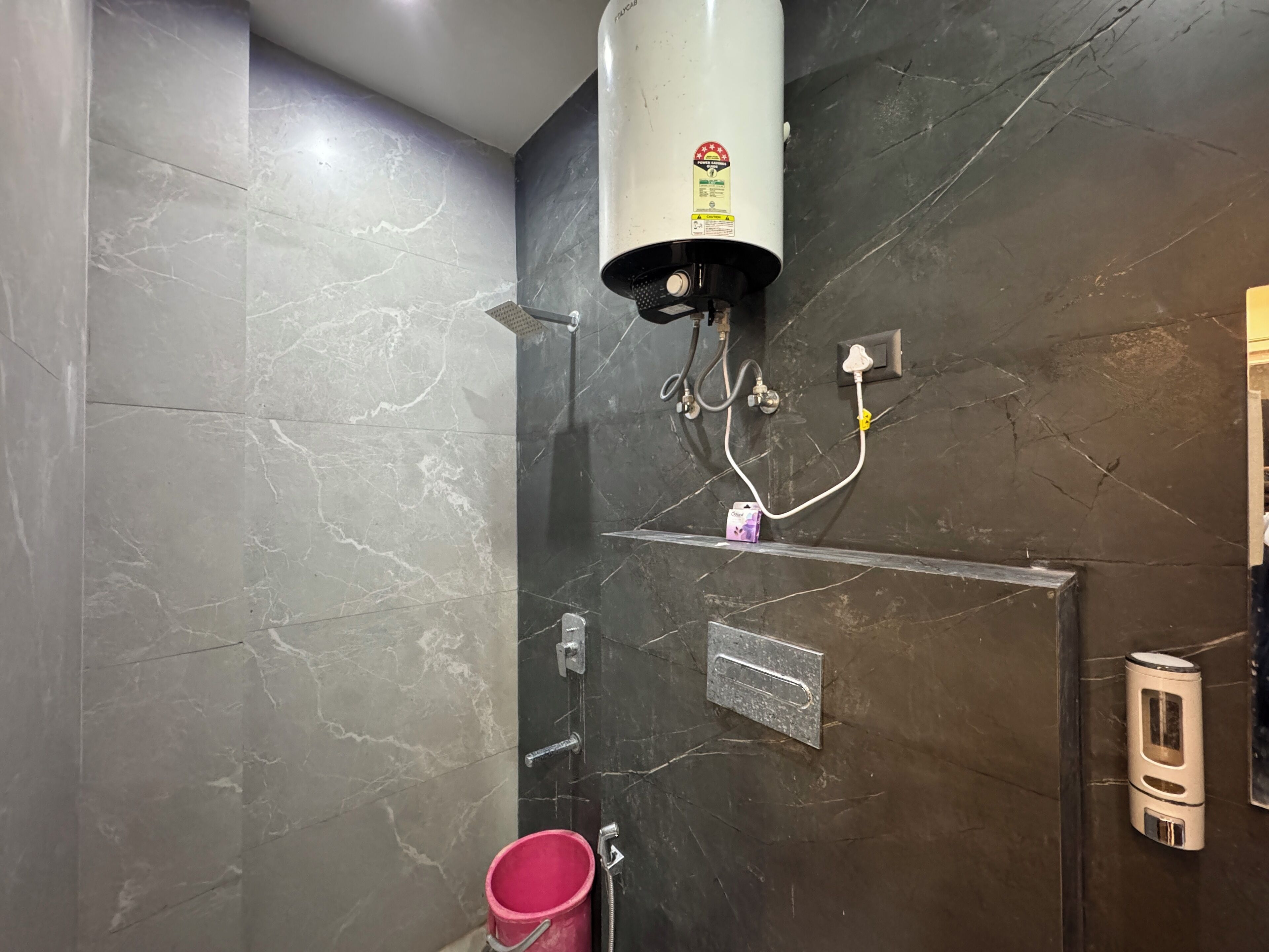 A bathroom with a water heater mounted on a dark marble wall, a pink bucket, and a soap dispenser.