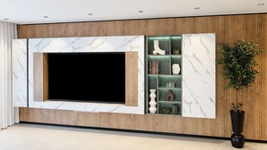 A modern living room with a large flat-screen TV mounted on a marble wall, wooden paneling, and a built-in shelving unit with decorative items. - Artemis (Tiberias)