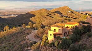 A hillside property with a winding road, a modern house with a flat roof, and a view of distant mountains and a power line tower. - Cozy villa with infinity pool in the mountains of Sierra Cabrera (TURRE)