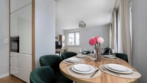 Comedor con una mesa redonda de madera dispuesta para cuatro personas, rodeado de sillas tapizadas de color verde. Un jarrón con flores rosas se encuentra sobre la mesa, y se observa una cocina al fondo.