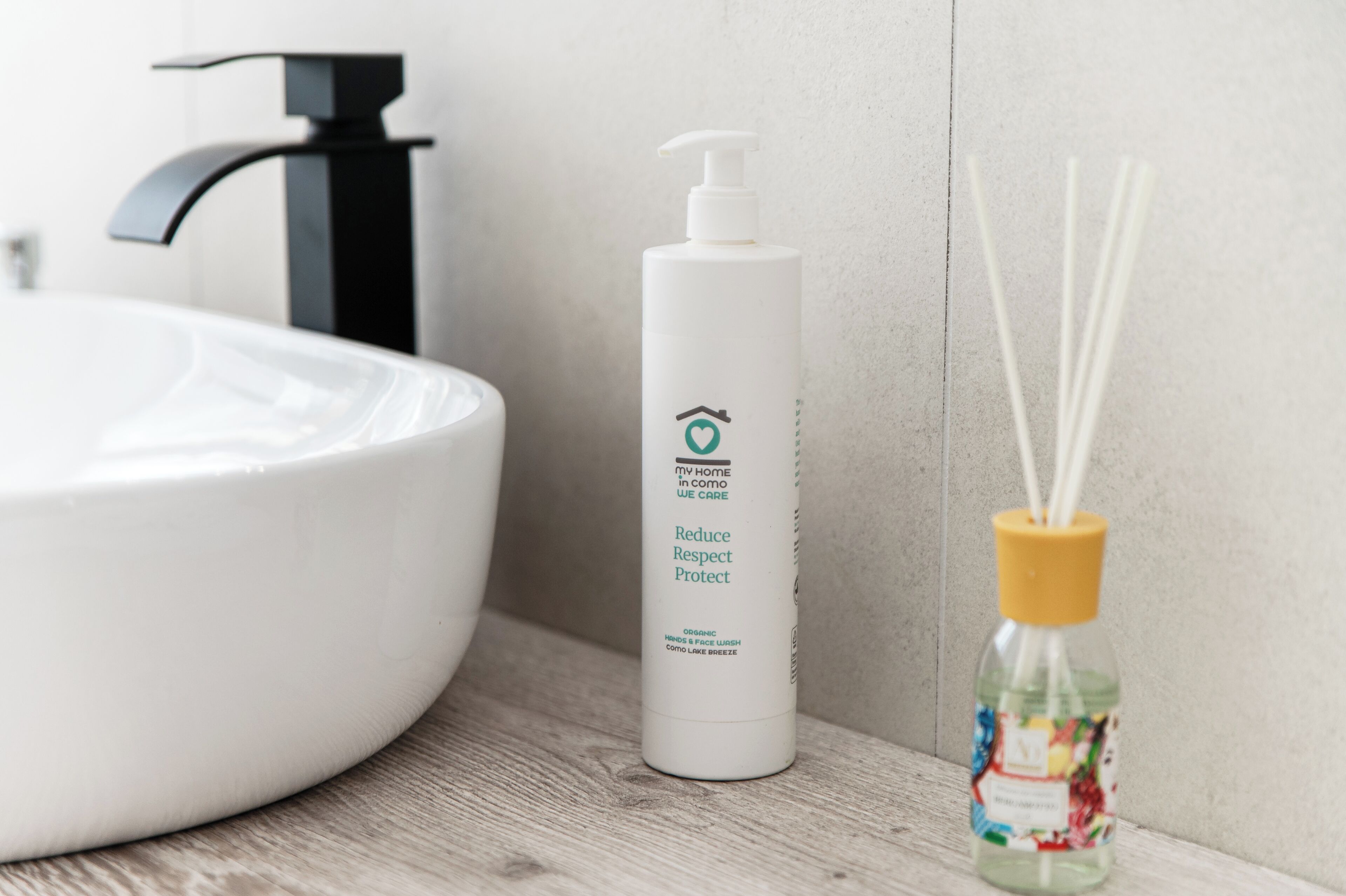 Un baño con un lavamanos blanco, una botella blanca de limpiador orgánico para manos y rostro, y un difusor de aroma de juncos.