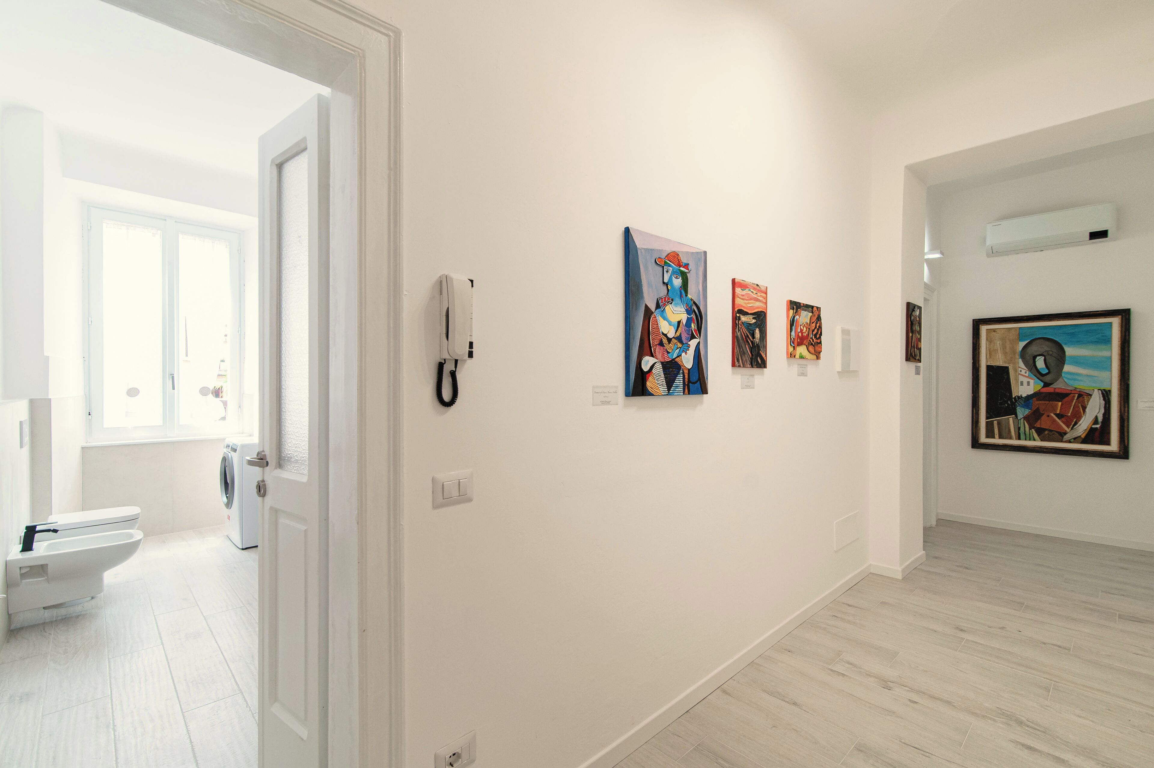 A gallery with artworks on the walls, a white door leading to a bathroom, and a light-colored floor.