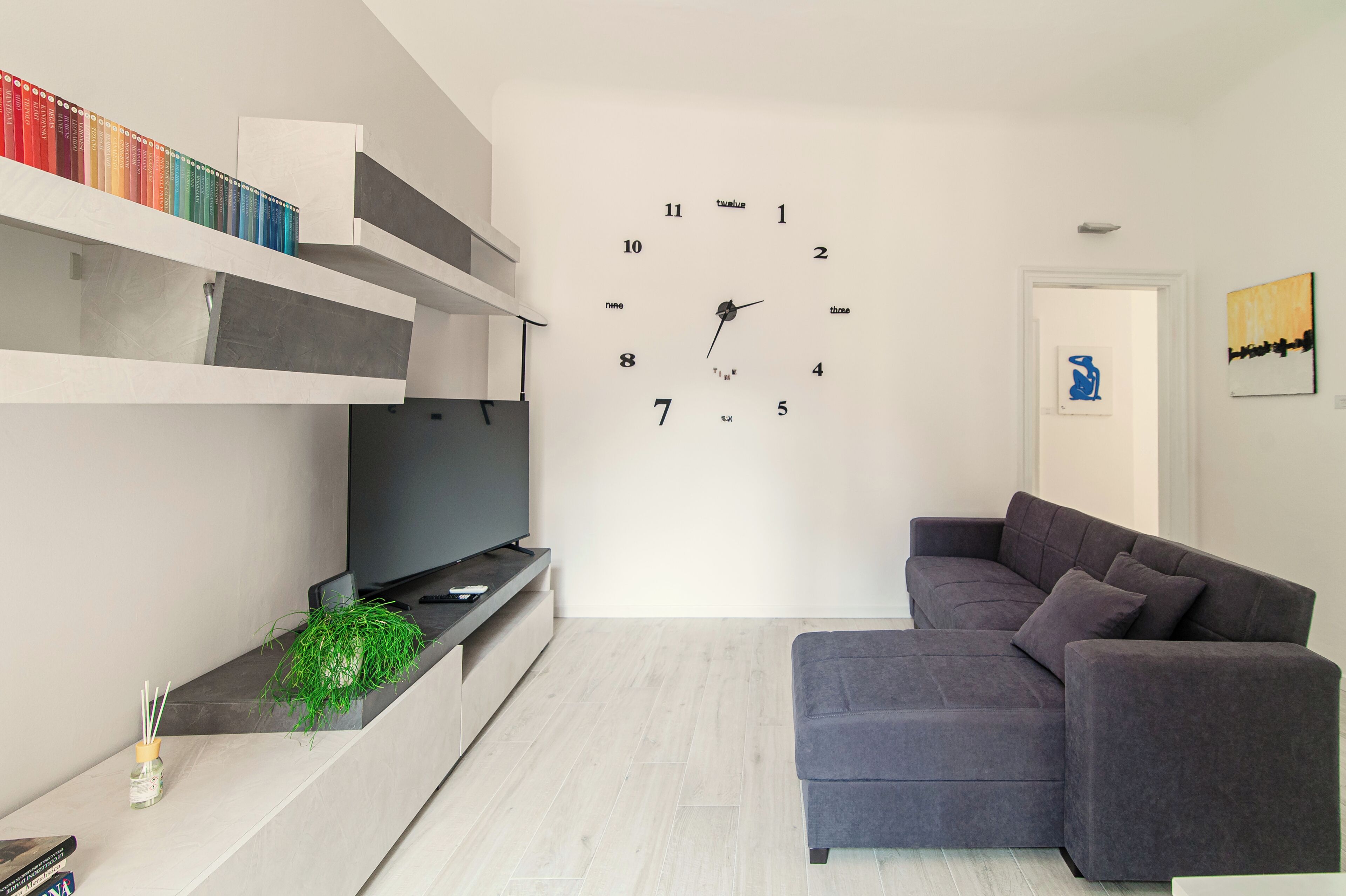 Ein modernes Wohnzimmer mit einer grauen Eckcouch, einem Flachbildfernseher an einer eingebauten Ablage, einer Wanduhr und einem Bücherregal mit farbenfrohen Büchern.
