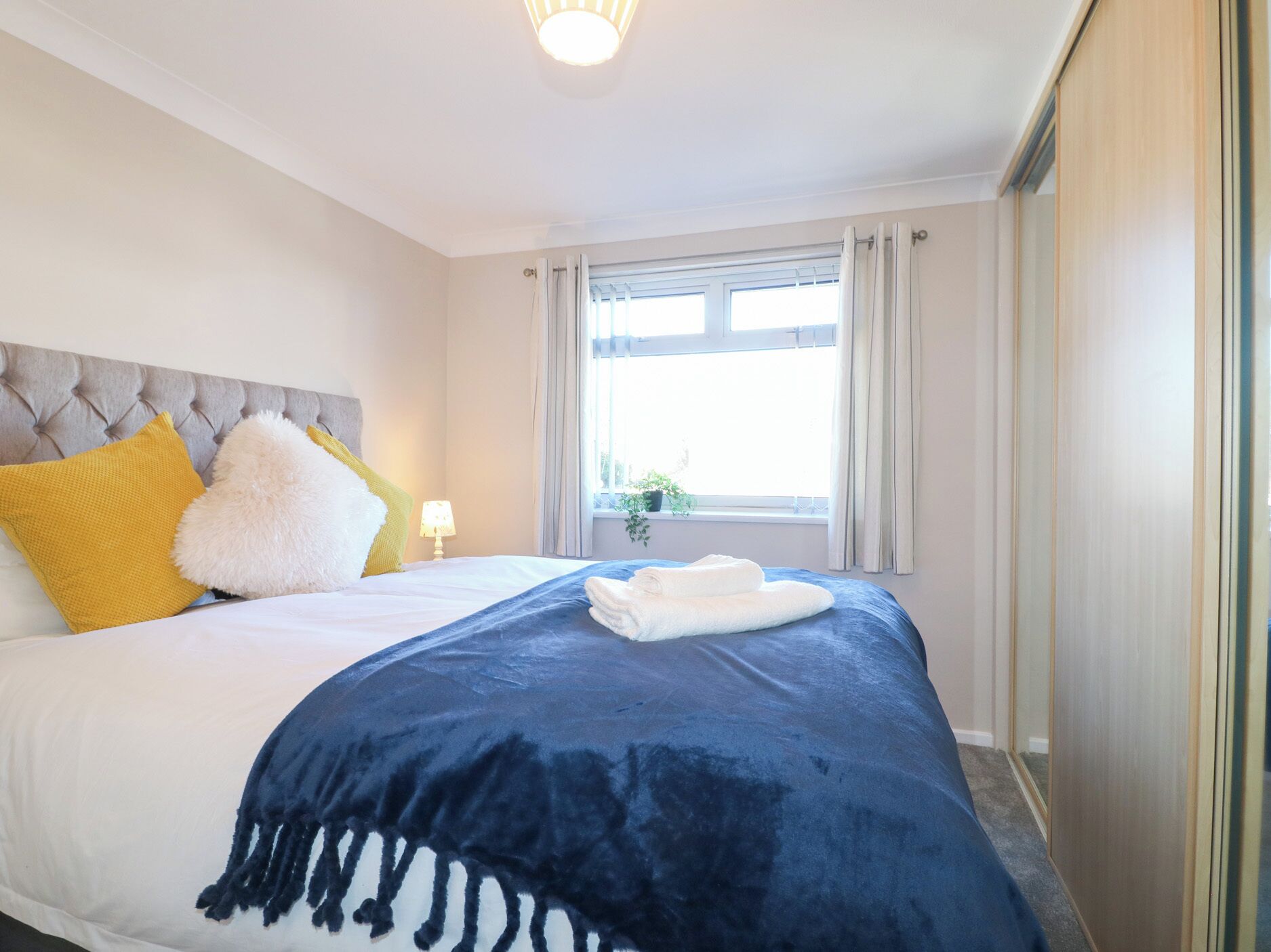 Une chambre d’hôtel avec un lit, une fenêtre avec des rideaux, une table de chevet avec une lampe et une plante en pot sur le rebord de la fenêtre.