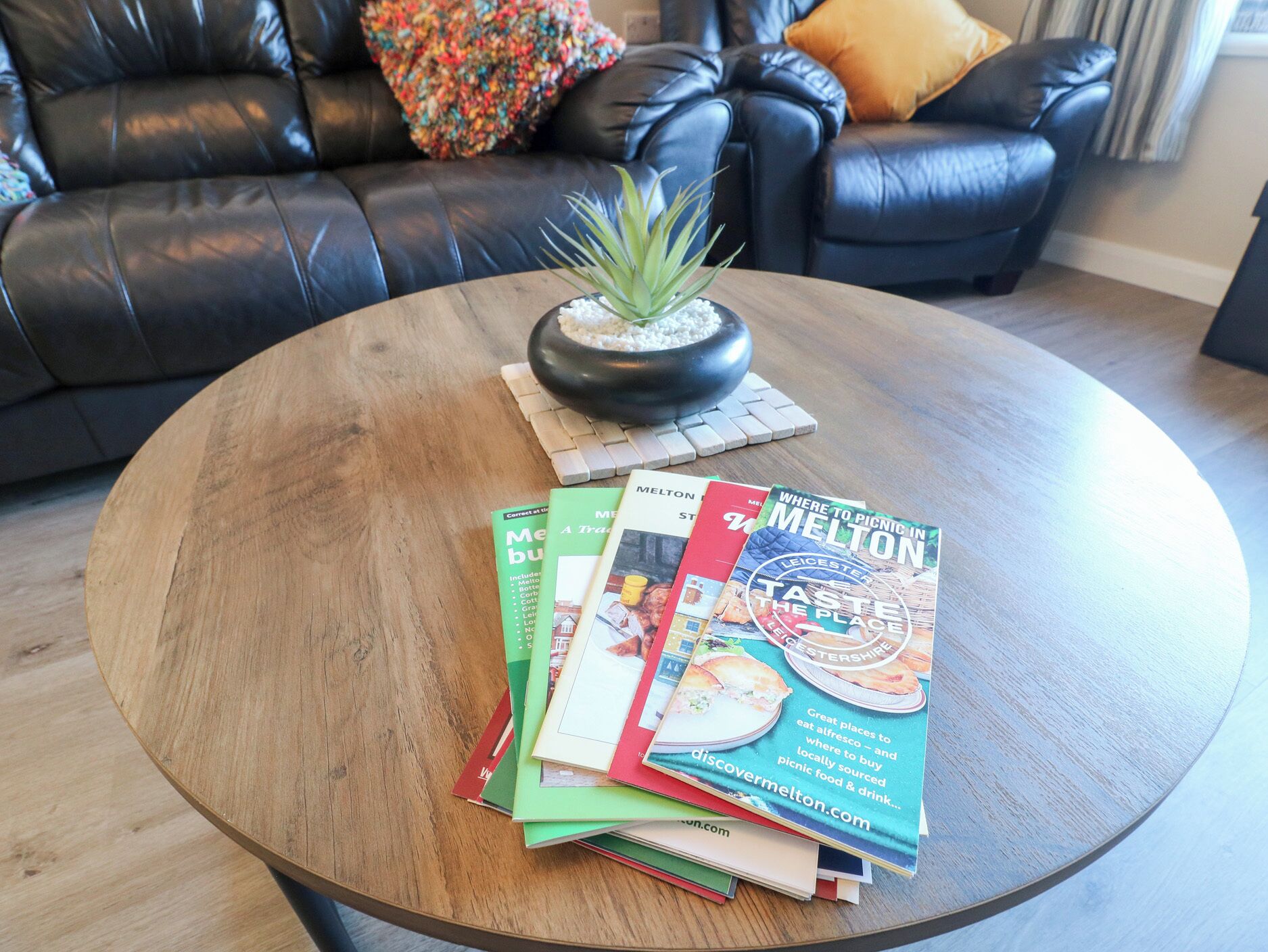 Uma mesa de centro redonda de madeira, com uma planta em vaso e uma pilha de revistas de viagem.