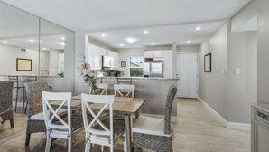 A modern kitchen with white cabinets, a central island, and a dining area with wicker chairs and a wooden table. - Gulf-Front Escape- 2BR Beachfront Condo by RVA (Longboat Key)