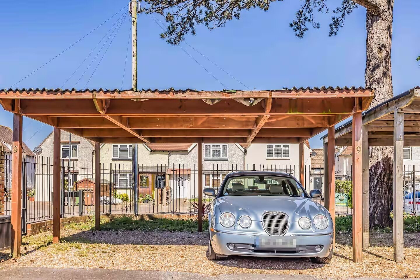 Un coche antiguo estacionado bajo una estructura de madera con una valla metálica y casas al fondo.