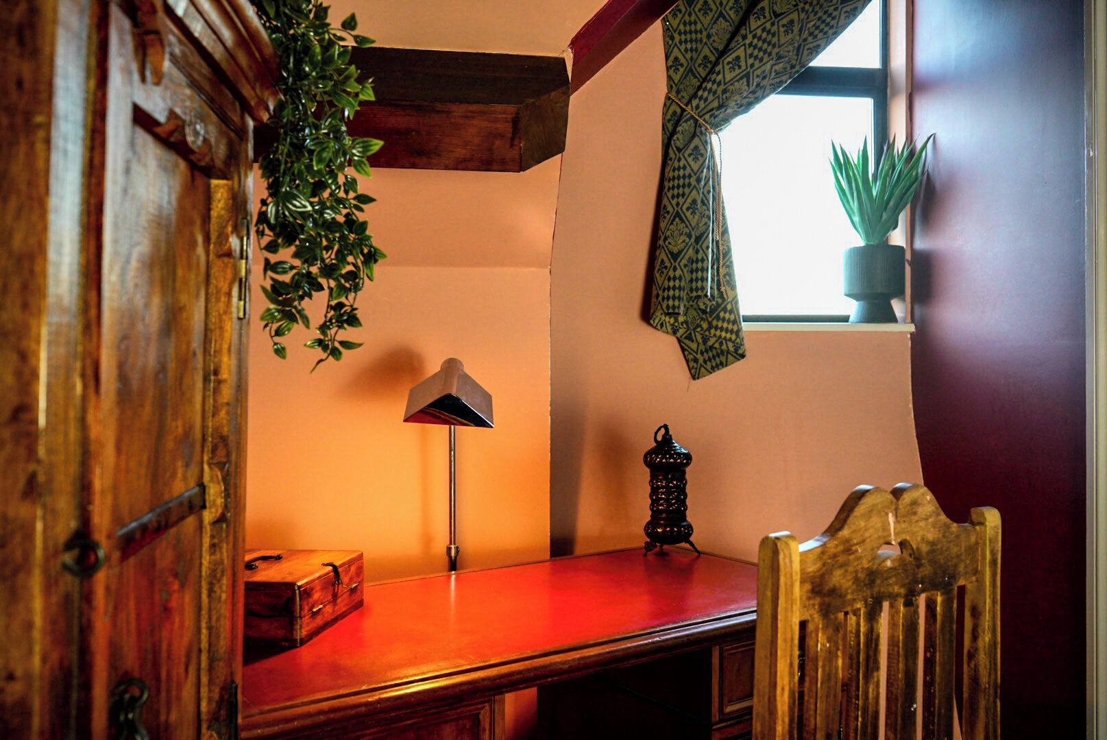 A corner room with a wooden desk, a lamp, a plant, and a window with patterned curtains.
