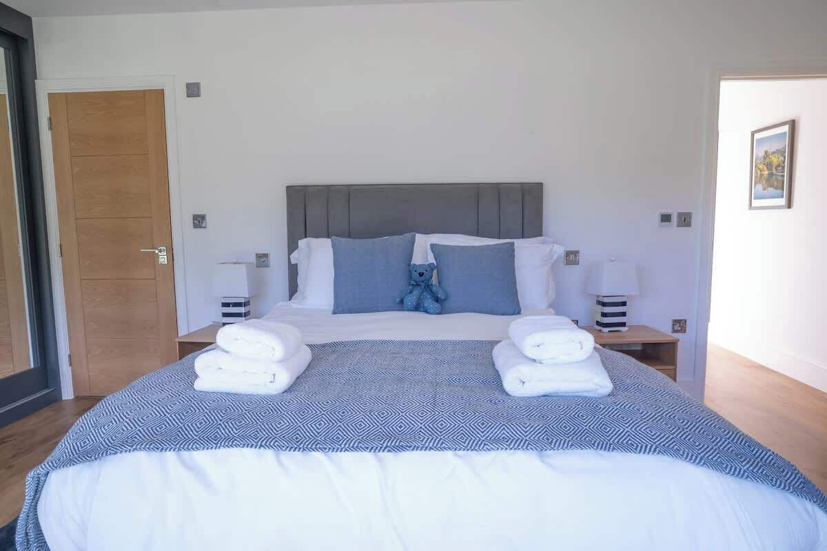Uma cama bem arrumada com cabeceira cinza, duas mesas de cabeceira com abajures e um cobreleito estampado em azul e branco.