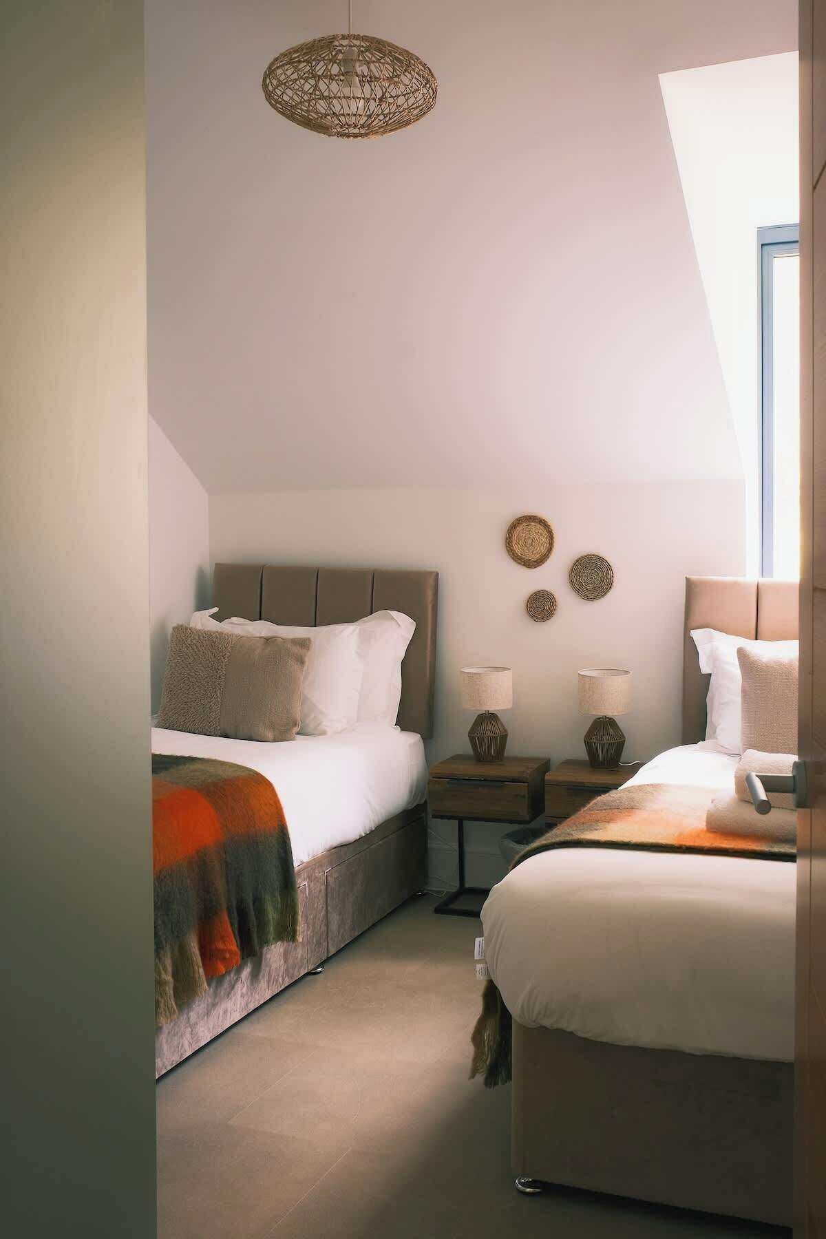 Une chambre avec deux lits, un luminaire suspendu, des tables de chevet avec des lampes et une vue sur l’extérieur par une fenêtre.