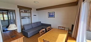 Una habitación con sofá, una mesa de comedor, sillas, lámpara, un cuadro en la pared y una ventana con cortinas.