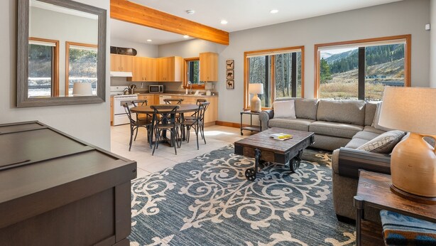 A modern living room with a sofa, coffee table, and a dining area with chairs. There is a kitchen in the background with wooden cabinets and a large window. - Ski Inn Condo 1 Bedroom Condo (Winter Park)