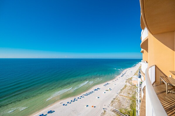 Uitzicht op het strand vanaf een balkon, met helderblauw water en een zandstrand, en parasols.