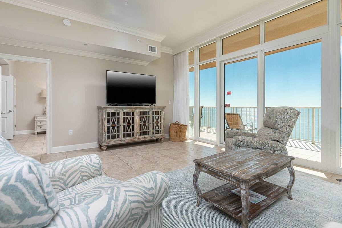A living room with a large TV, a wooden coffee table, patterned sofas, and a balcony with a view.