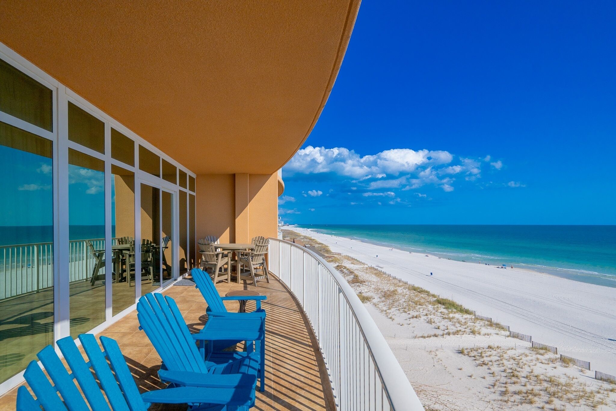 Un balcon avec des chaises bleues et une table donnant sur une plage où l’eau est d’un bleu limpide et le sable est blanc.