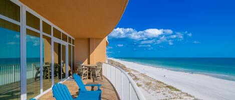 Un balcon avec des chaises bleues et une table donnant sur une plage où l’eau est d’un bleu limpide et le sable est blanc.
