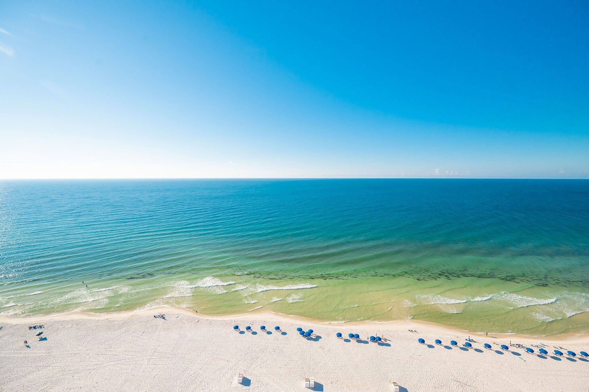 An aerial view of a beach with clear blue water, a sandy shore, and rows of blue umbrellas.