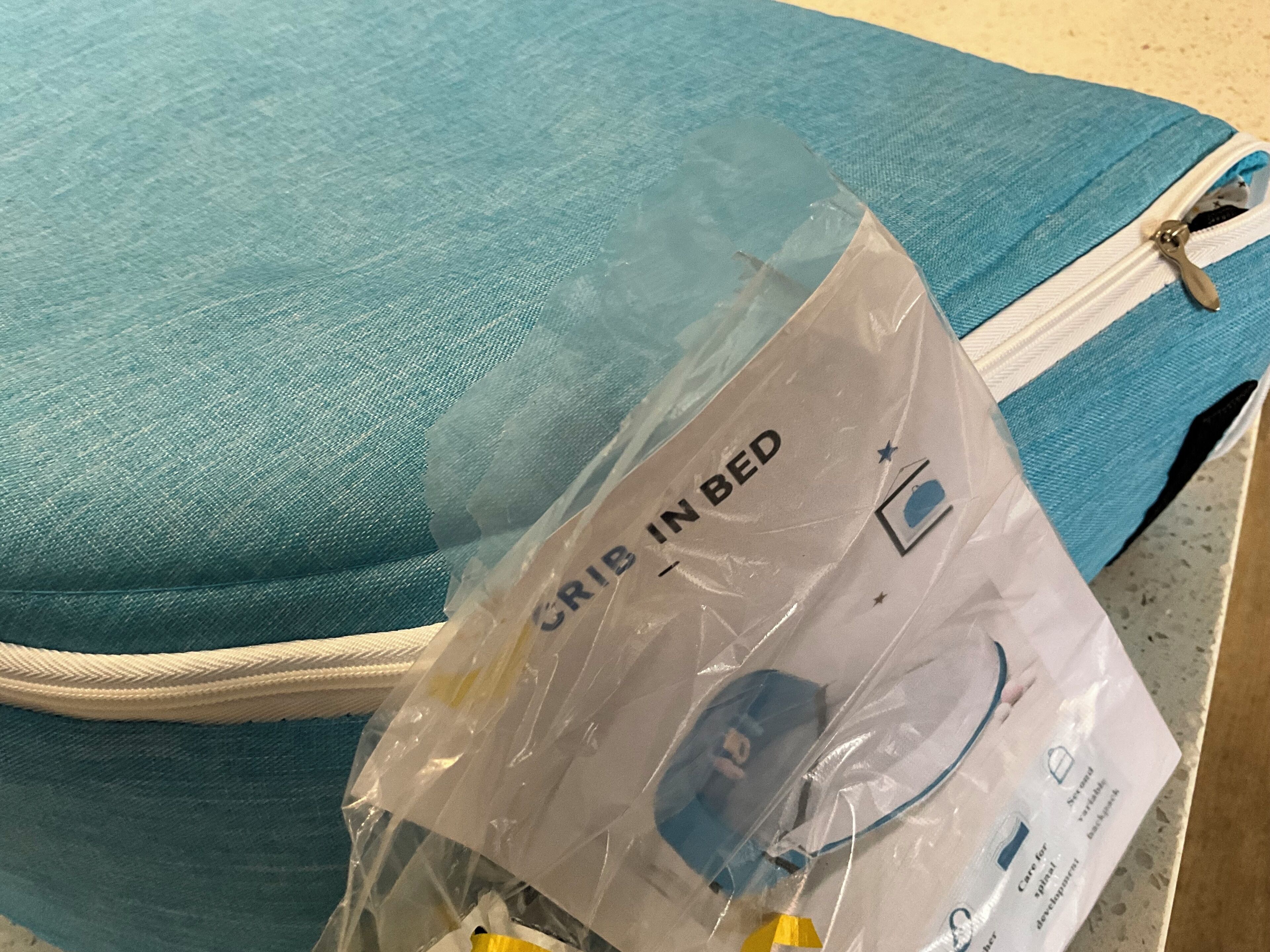 Een blauw sprei met een zak met rits en een plastic zakje met de tekst 'BEDDITJE IN BED' dat een babybedje bevat.