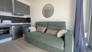 Ein kompakter Wohnbereich mit Küchenzeile, einem grünen Sofa mit Kissen, einem runden Spiegel und einem fenster mit Vorhängen.