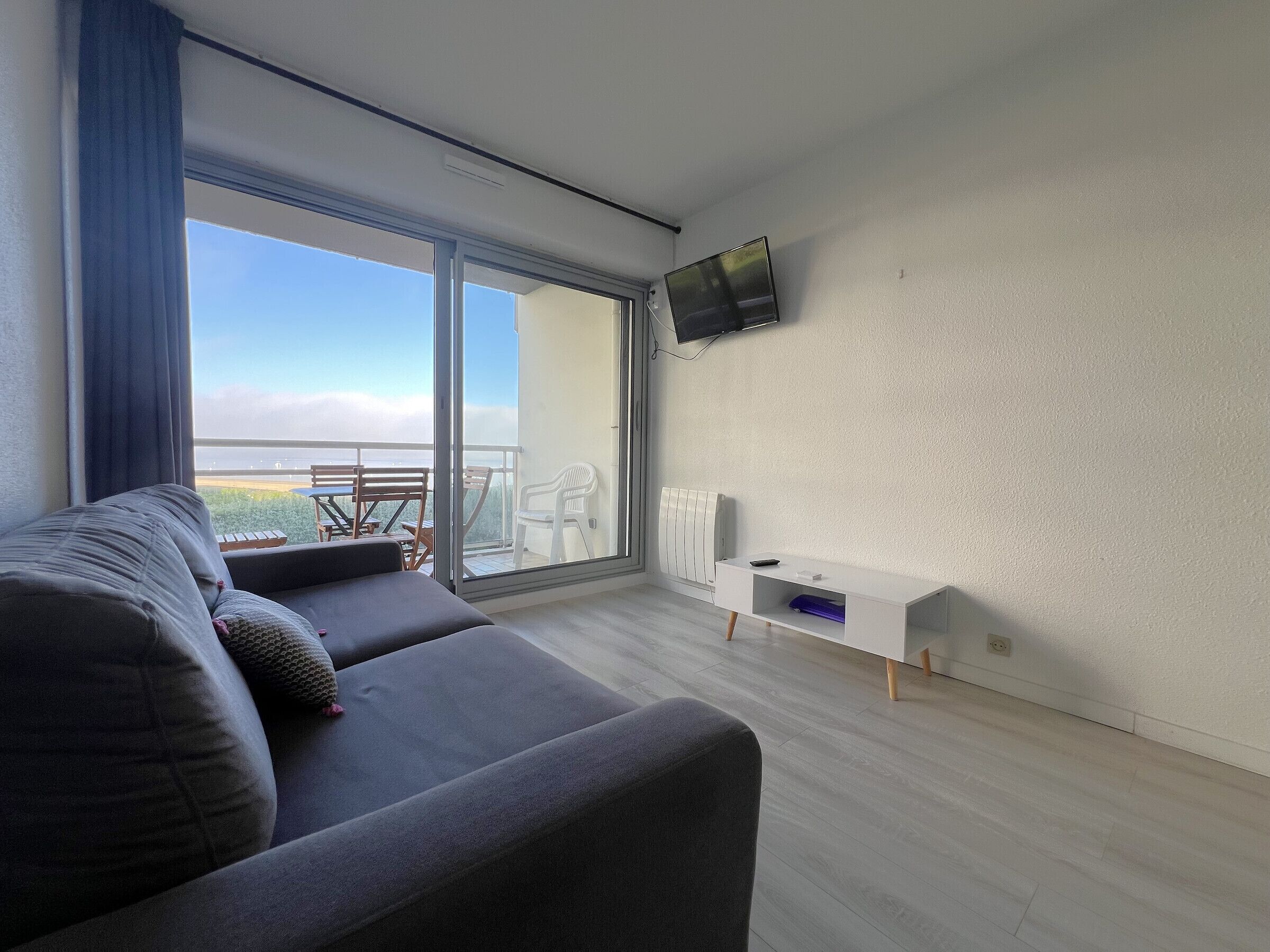 Un salón moderno con un sofá gris, un televisor de pantalla plana montado en la pared y un balcón con vistas al mar.