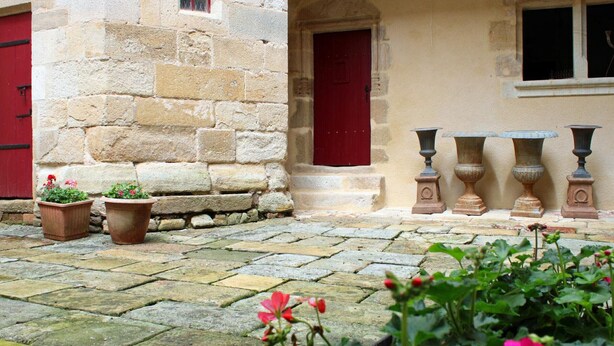 Un edificio de piedra con puertas y ventanas rojas, plantas en macetas y jardineras de piedra en un camino adoquinado.