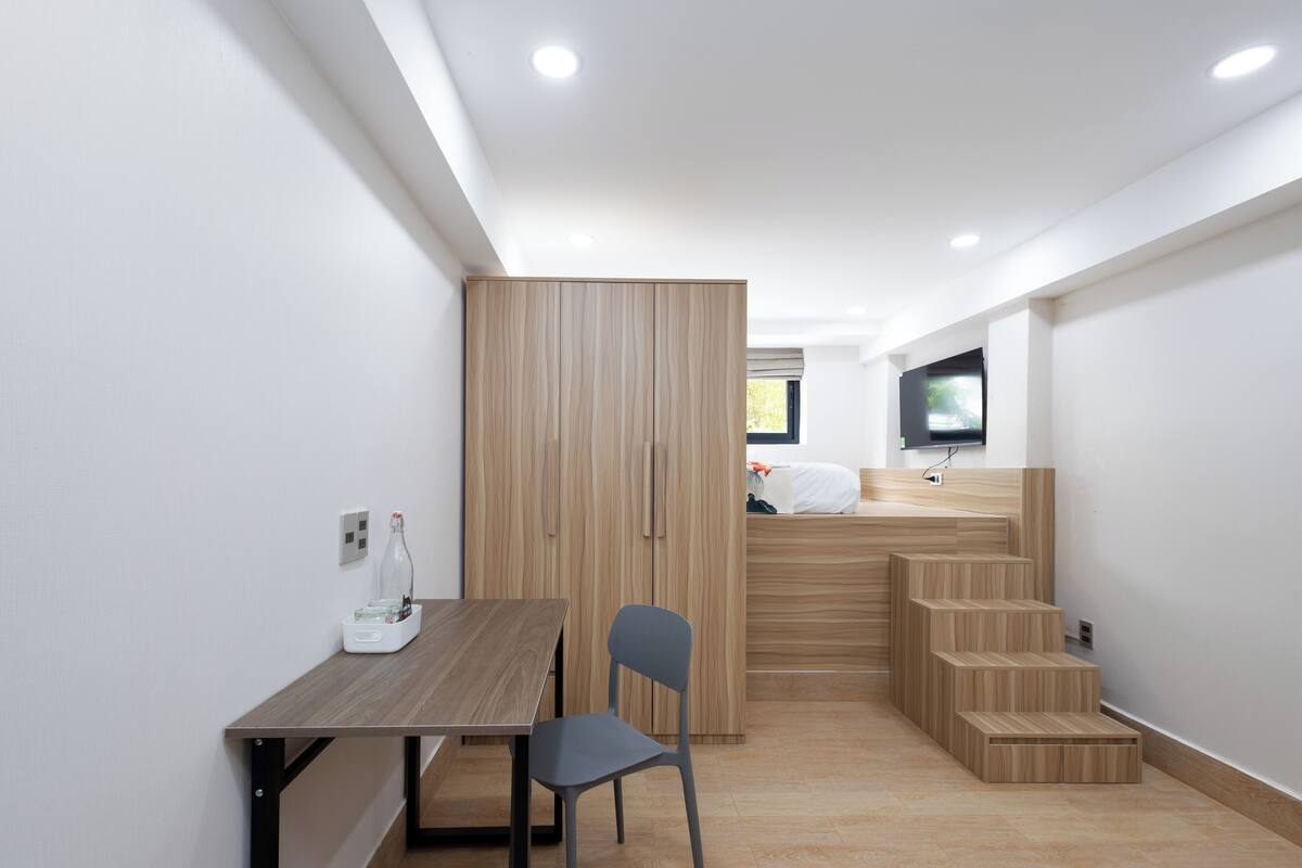 Ein kleines Zimmer mit einem Holztisch, einem Stuhl, einem Kleiderschrank und einem an der Wand befestigten Fernseher.