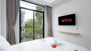 Une chambre d’hôtel dotée d’une grande fenêtre, d’un lit recouvert d’une couette blanche, d’un téléviseur à écran plat fixé au mur et d’une étagère en bois au-dessus du lit.