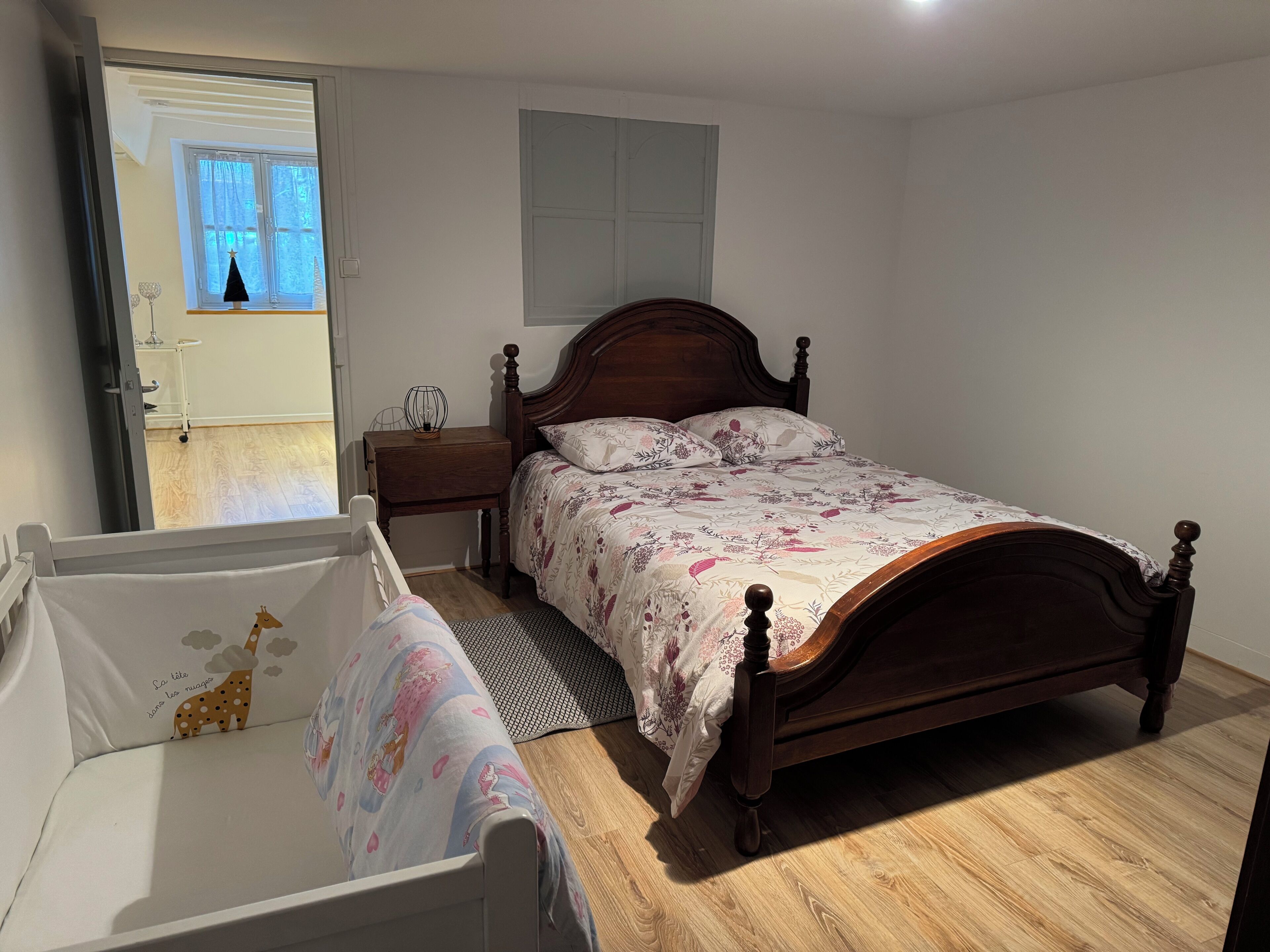 A bedroom with a wooden bed, a nightstand, a crib, and a window with curtains.