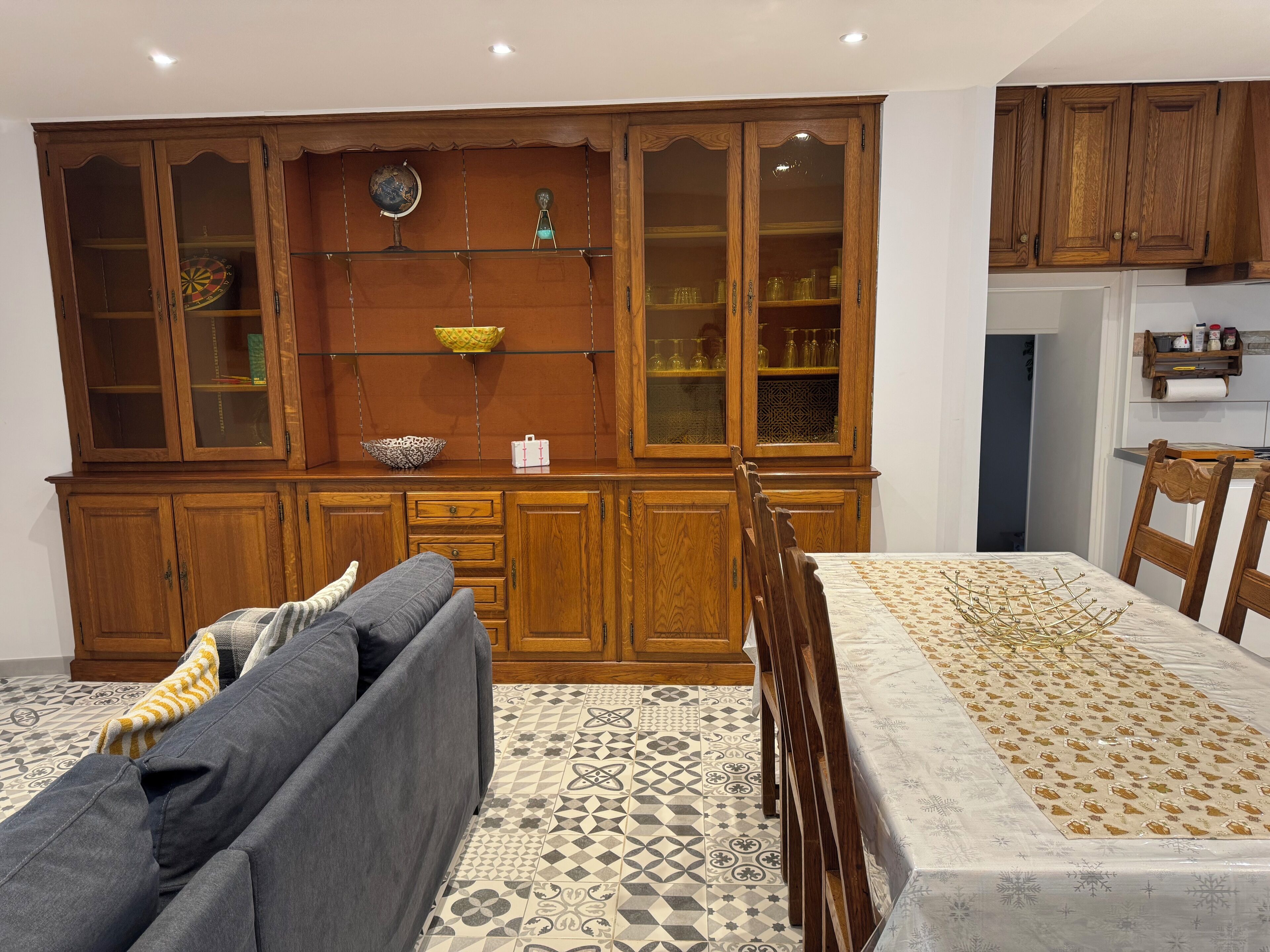 A dining area with a wooden cabinet, a table with a patterned tablecloth, and a sofa with cushions.