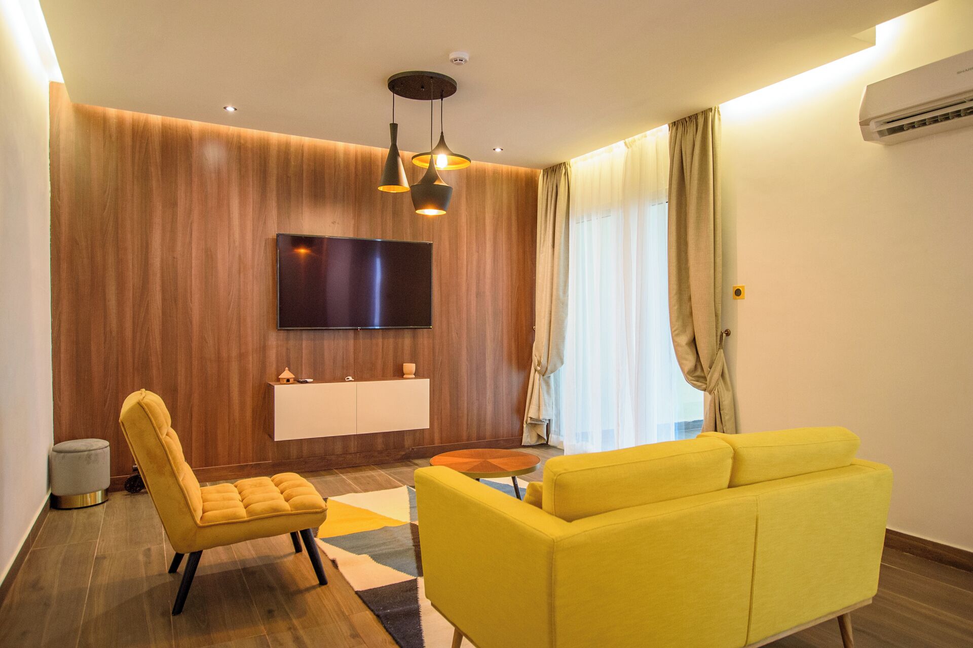 A modern living room with a yellow sofa, a yellow armchair, a wooden accent wall, a flat-screen TV, and a small round table.