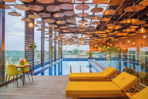 A modern indoor pool area with a view of the city, yellow lounge chairs, and a ceiling with circular light fixtures.