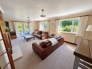 A spacious living room with a brown leather sofa, a coffee table, and a fireplace. There are large windows providing natural light and a view of greenery outside. - Cae Erwyn - Coastal Escape with Hot Tub, Cinema & Games Room (Anglesey)