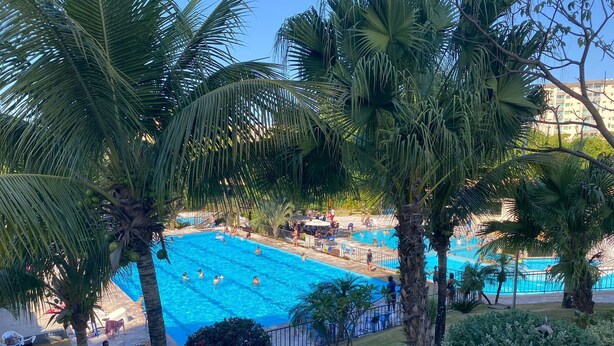 A swimming pool surrounded by palm trees and a grassy area with a fence. - Modern apartment with complete leisure area and access to the Golden Dolphin. (Caldas Novas)